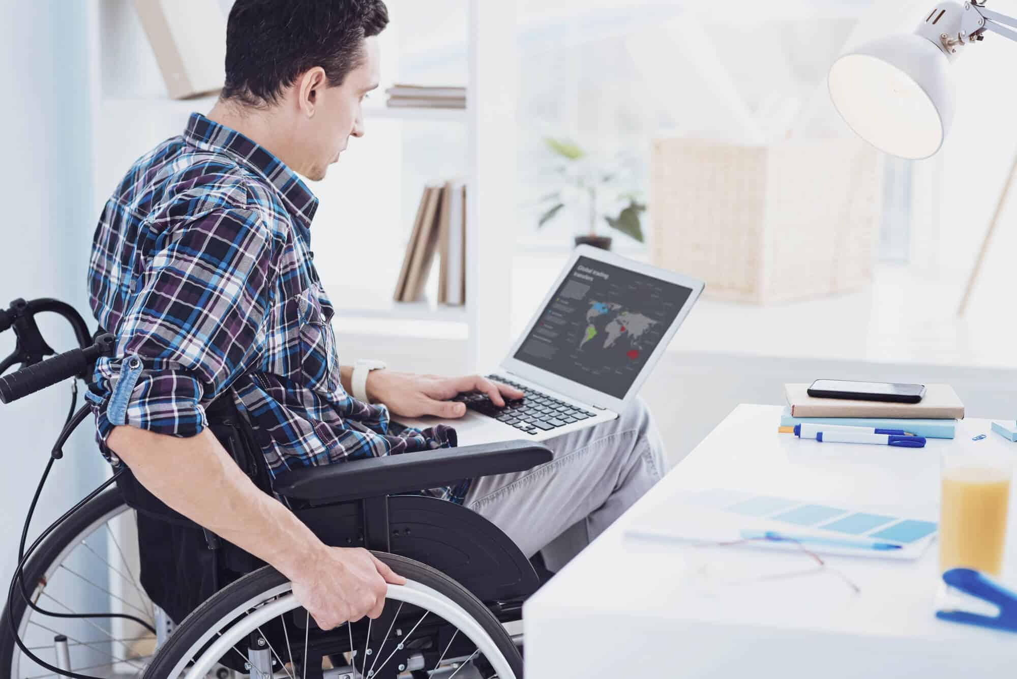 A person in a wheelchair utilizes a laptop at a white desk with office supplies, highlighting the importance of ADA compliance in creating accessible workspaces.