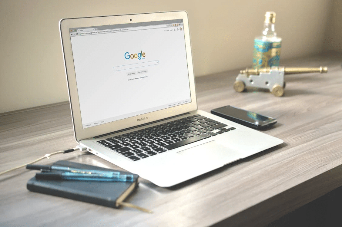 A laptop displaying Google's homepage is on a wooden desk next to a smartphone, a notebook with pens, and a decorative cannon with a small glass bottle.