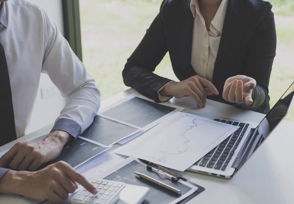 Two people in business attire sit at a desk with a laptop, calculator, charts, and papers, discussing financial data and reviewing client SEO reports.