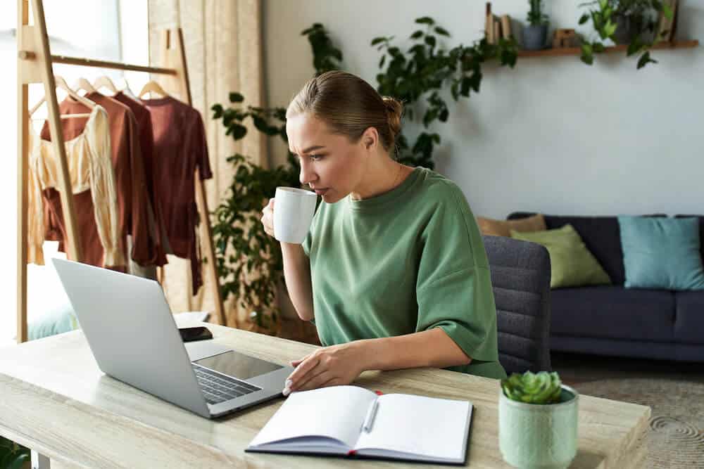 Woman sitting at a desk, drinking from a white mug while looking at a laptop, with an open notebook and pen in front of her—perfecting client SEO reports in a cozy home office setting.