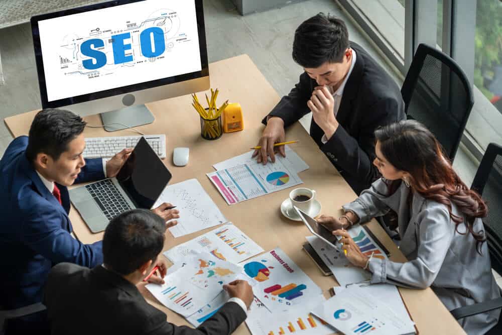 Four business professionals review charts and graphs around a table, with a computer displaying "SEO" in the background, highlighting their focus on automated SEO reporting to enhance performance tracking.